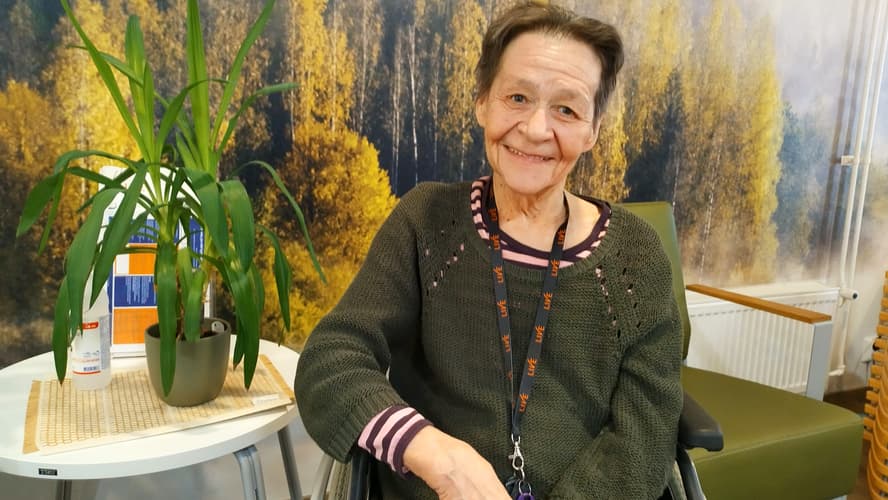 Helena Tervonen istuu pyörätuolissa hymyillen. Metsäinen seinä taustalla.  featured-image