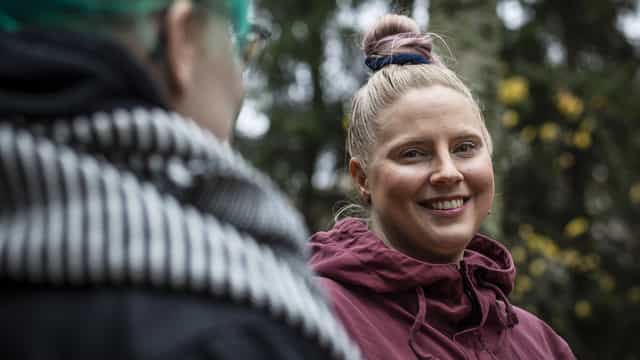 Henkilö hymyilee metsässä. Etualalla toinen henkilö selkäpäin. Lähikuva. 
